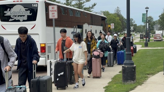 Many people walk towards the viewer, away from a large white bus, and carry luggage with them